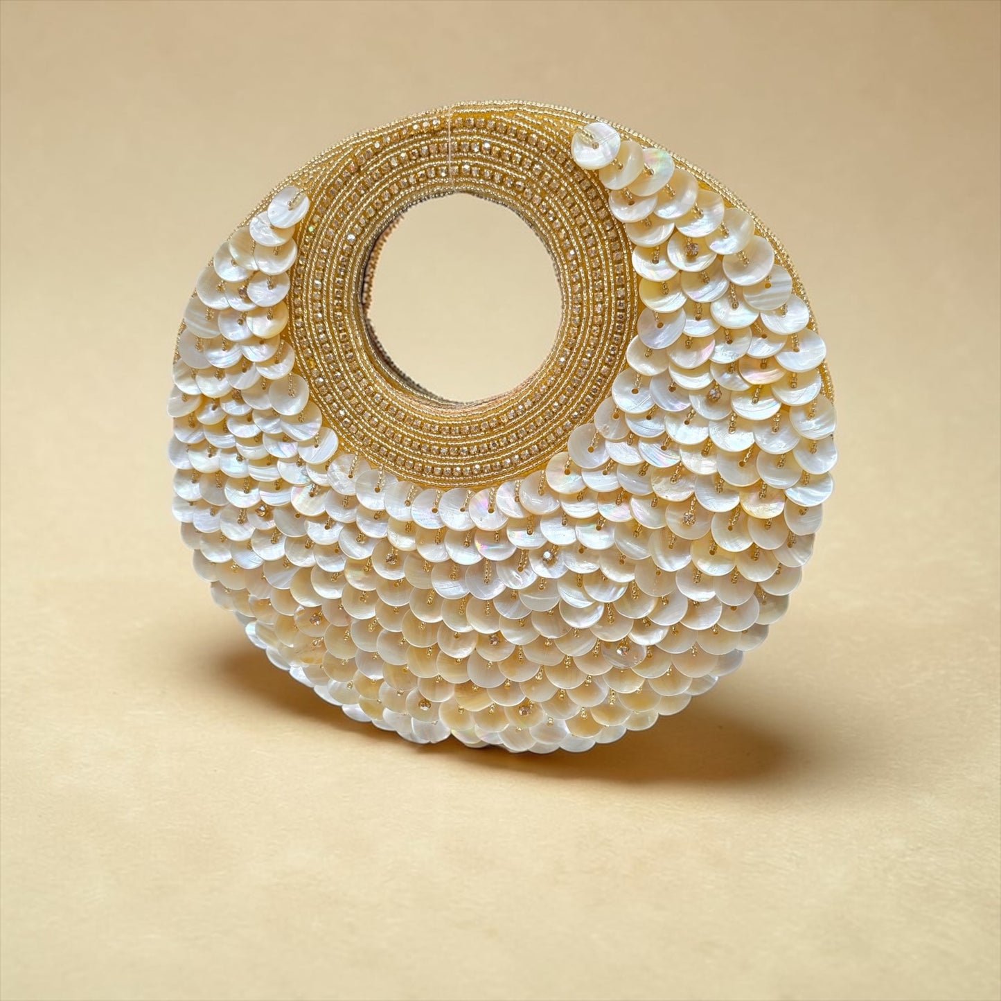 Dual-sided hand-embroidered circular mother of pearl clutch featuring a central floral shell motif with diamond-cut crystal accents and premium seed pearls. This 8.5-inch slim-profile wedding bag is crafted on a natural jute base with a hidden magnetic closure, perfect for luxury bridal and ethnic festive wear