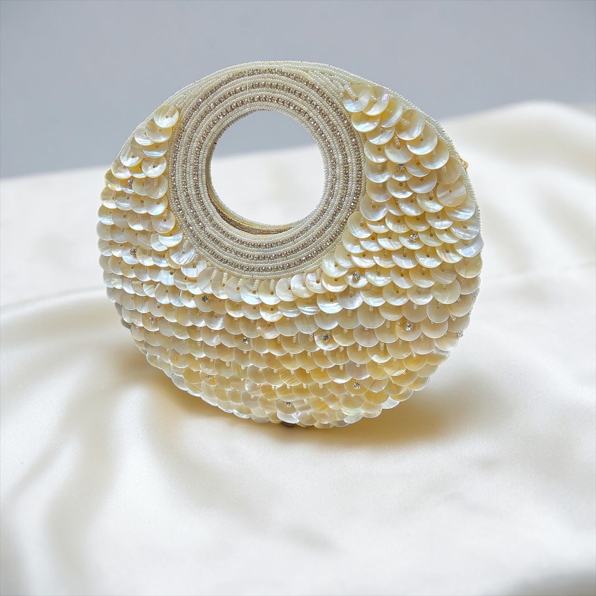 Dual-sided hand-embroidered circular mother of pearl clutch featuring a central floral shell motif with diamond-cut crystal accents and premium seed pearls. This 8.5-inch slim-profile wedding bag is crafted on a natural jute base with a hidden magnetic closure, perfect for luxury bridal and ethnic festive wear