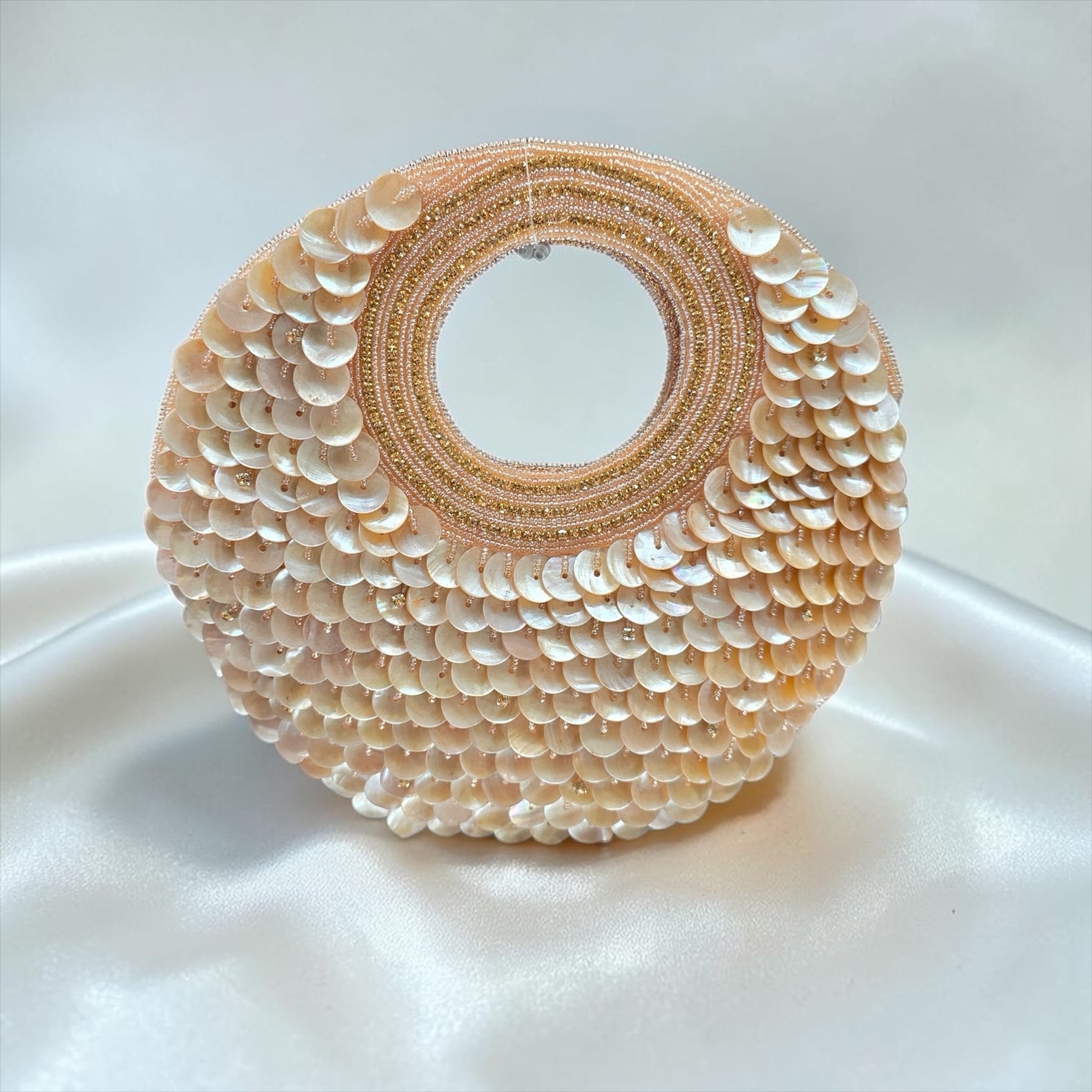 Dual-sided hand-embroidered circular mother of pearl clutch featuring a central floral shell motif with diamond-cut crystal accents and premium seed pearls. This 8.5-inch slim-profile wedding bag is crafted on a natural jute base with a hidden magnetic closure, perfect for luxury bridal and ethnic festive wear